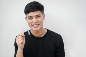 Young man wearing black sweater smiling and holding clear aligner.